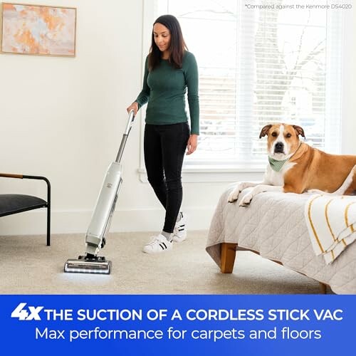 Woman using a cordless stick vacuum on carpet with dog on bed.